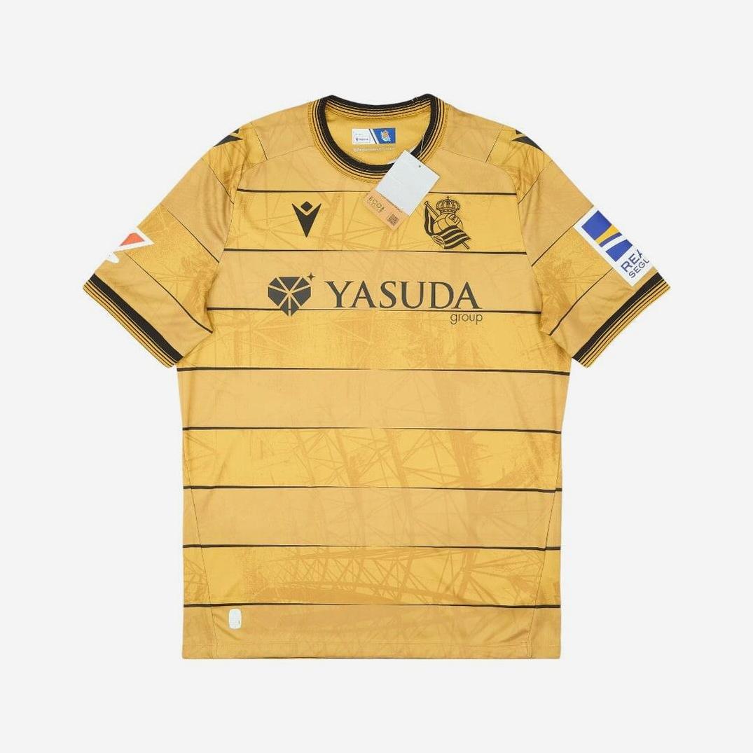 The Real Sociedad 2024-25 Away Shirt by Retro1999 features gold fabric, black horizontal stripes, a Yasuda Group sponsor logo, club crest on the chest, and sleeve patches—a standout La Liga jersey for any fan.