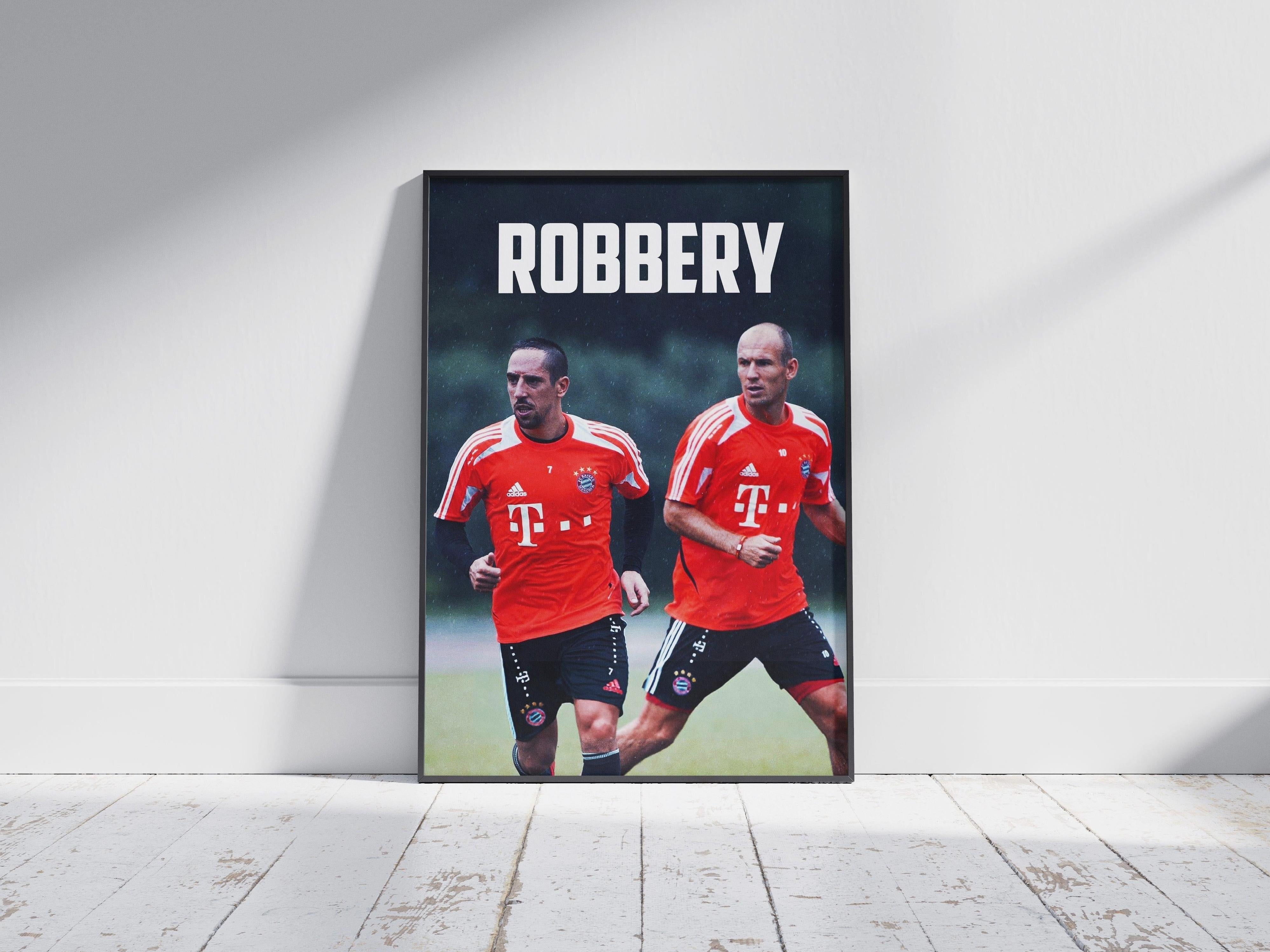 The Retro1999 Robben x Ribery ROBBERY Poster shows the duo in red Bayern Munich jerseys sprinting, with ROBBERY in bold above, framed and placed on a wooden floor as sunlight creates dramatic shadows on a white wall behind.