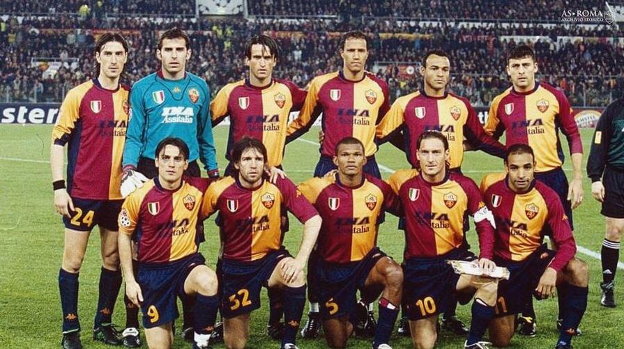 A soccer team in Retro1999s Roma 2001-02 Home Shirt, featuring red and yellow uniforms, poses on the field before a match with eleven players in two rows and a stadium crowd in the background.