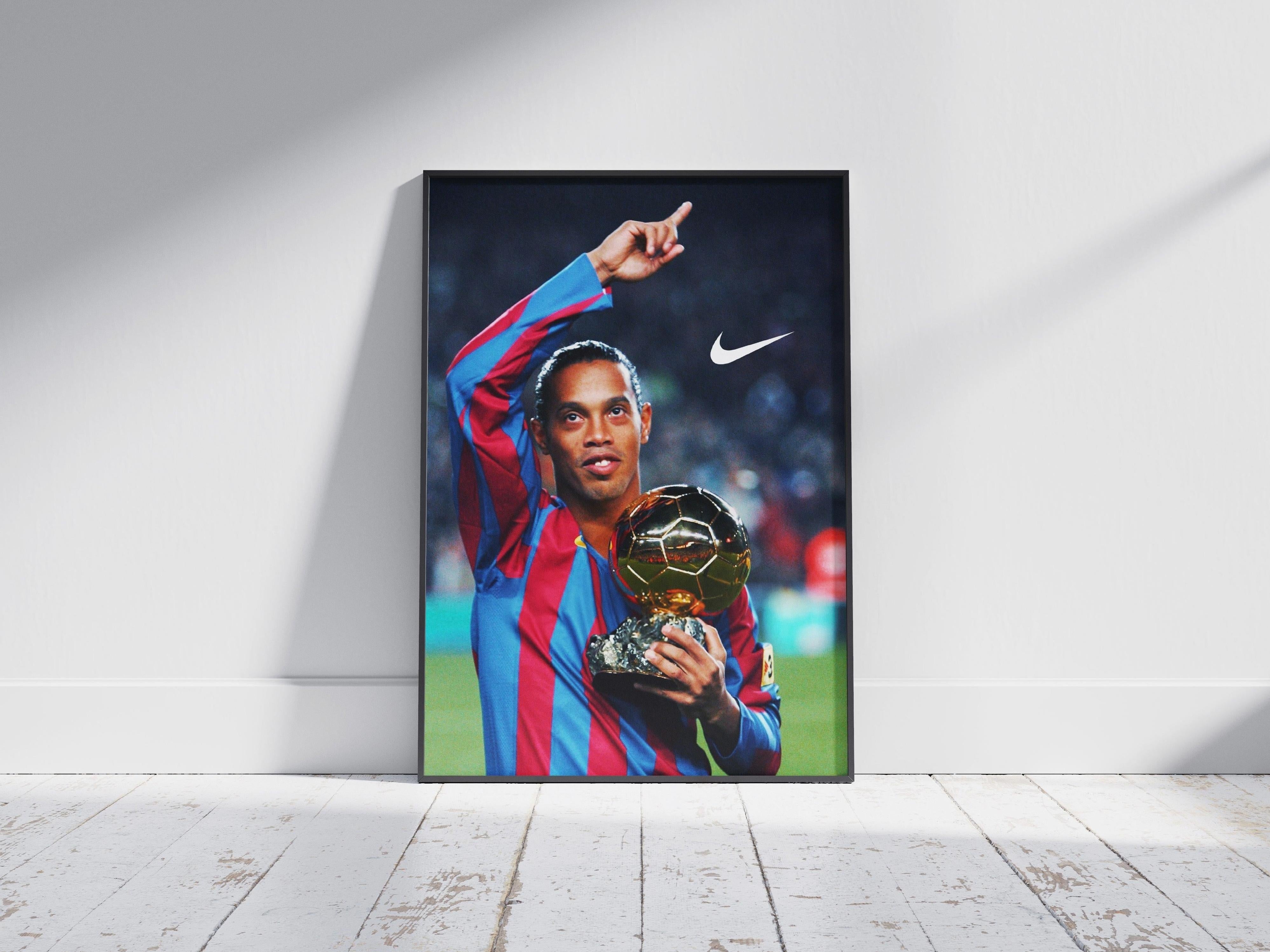 The Retro1999 Ronaldinho x Ballon dOr Poster features the football legend in a red and blue jersey holding a golden trophy, pointing upward, with a Nike logo in the top right corner.