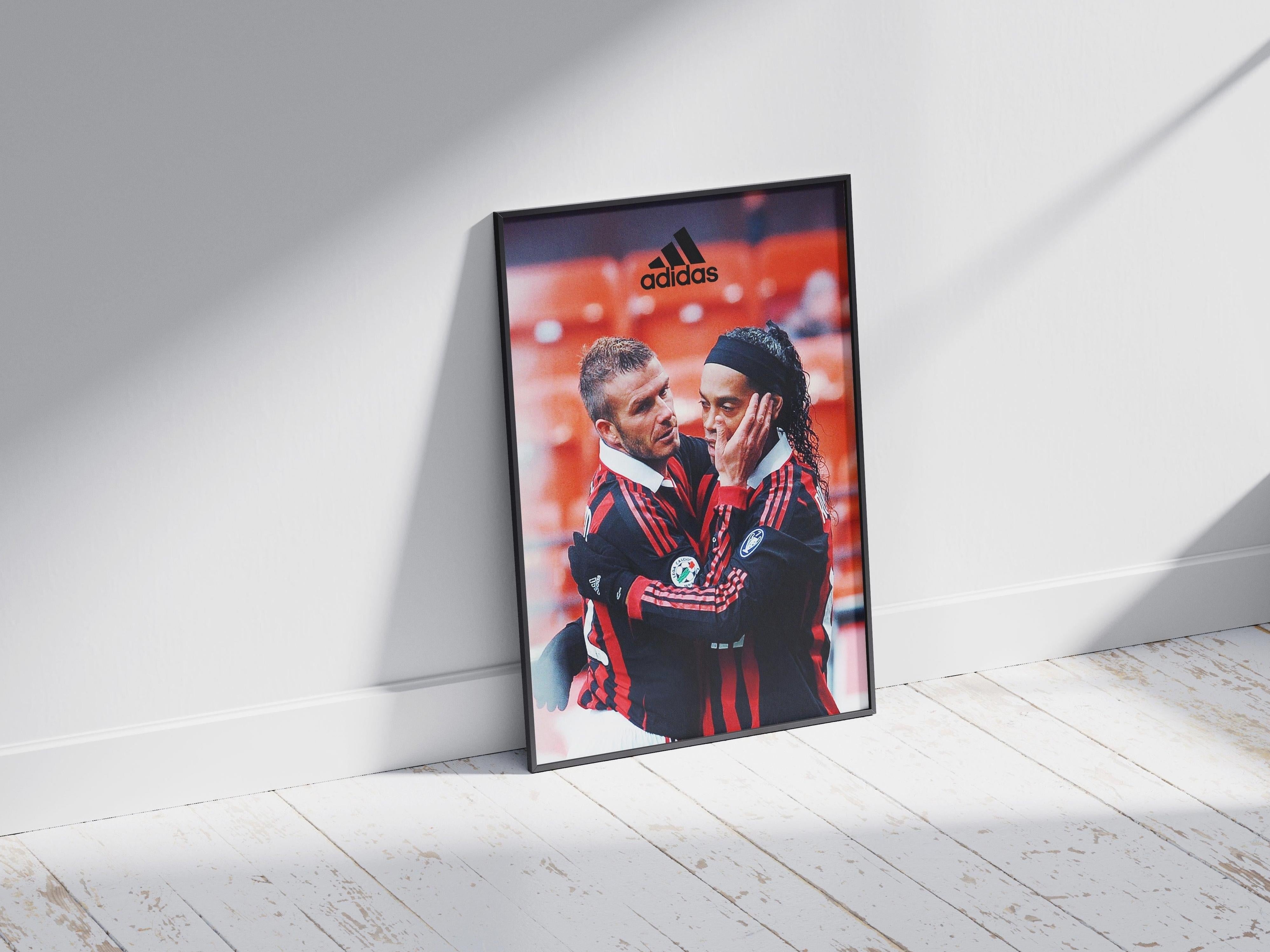 The Ronaldinho x David Beckham Poster by Retro1999 features two soccer legends in red and black jerseys embracing on the pitch, capturing a classic Milan Serie A moment—perfect memorabilia for football fans.