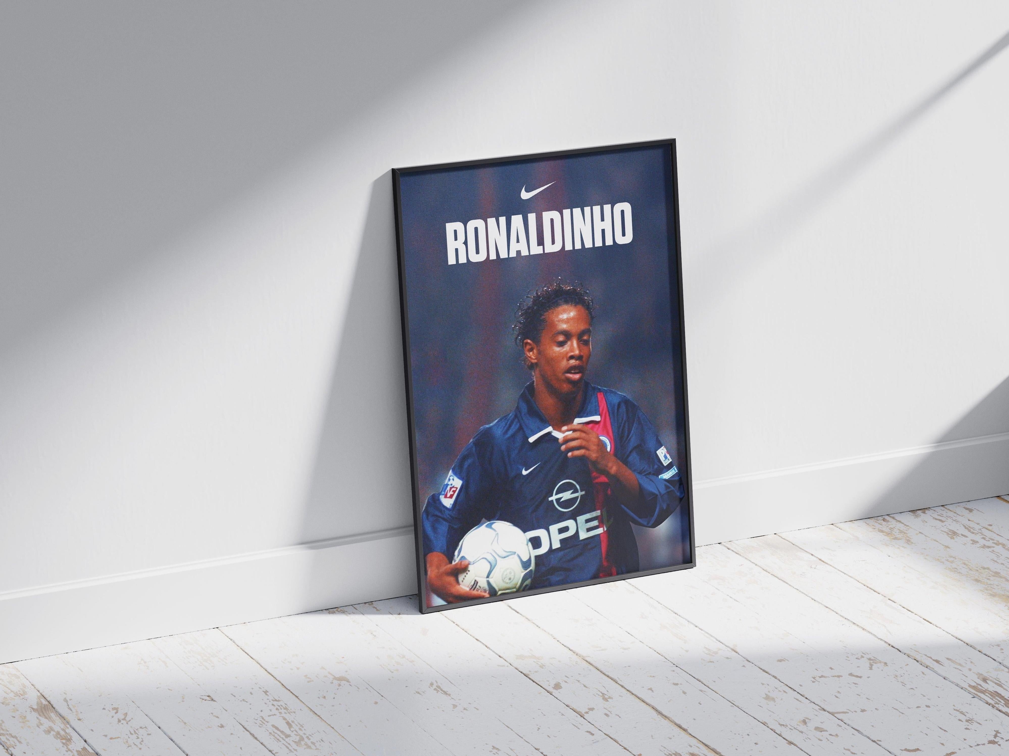 The Retro1999 Ronaldinho X PSG Poster shows Ronaldinho in a blue and red PSG jersey holding a ball, with his name and a Nike logo above—ideal football memorabilia for fans, displayed leaning on a white wall in bright sunlight.