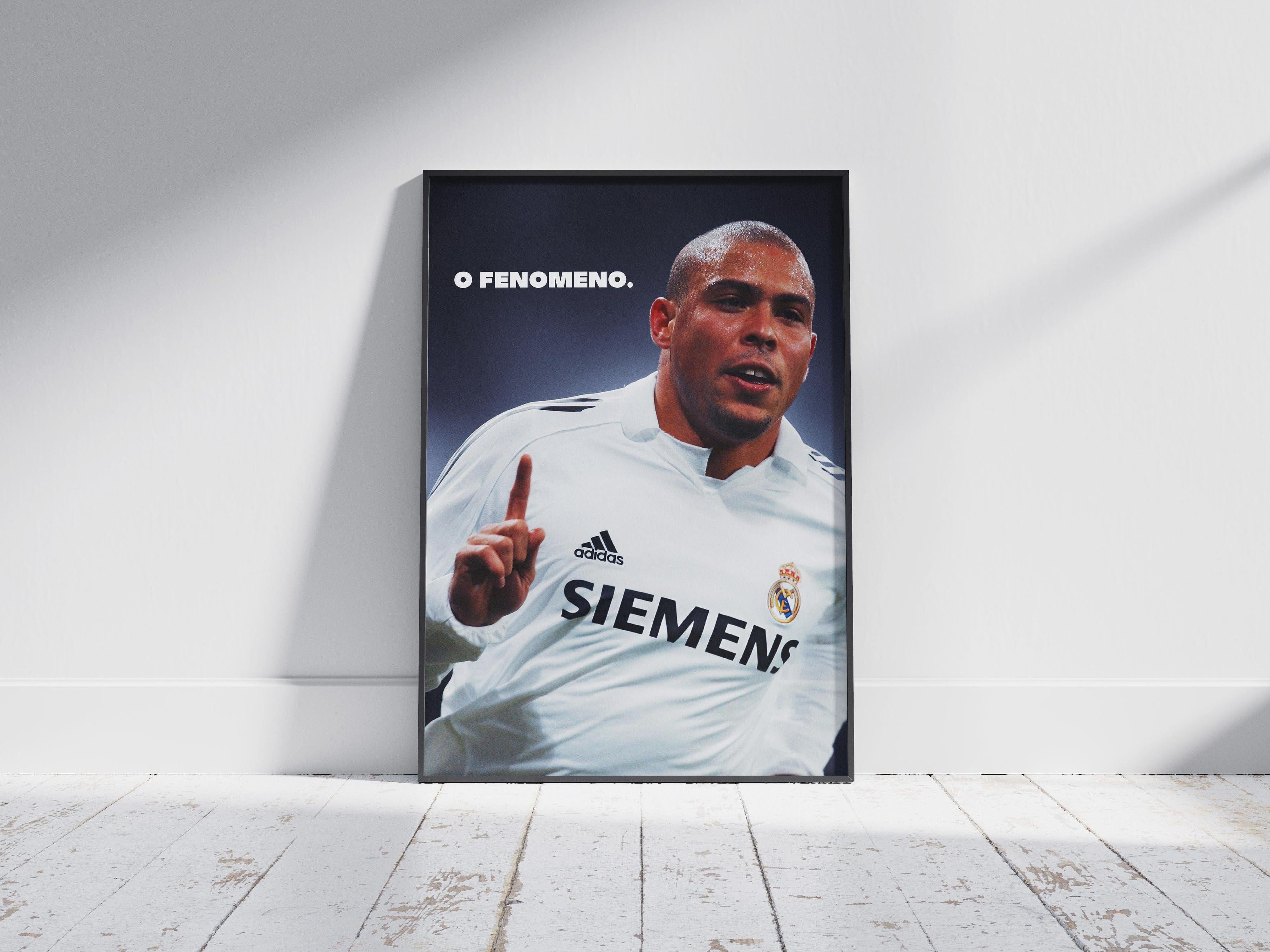 The Retro1999 Ronaldo O Fenômeno Poster shows Ronaldo in a white SIEMENS jersey, raising one finger. O FENOMENO is printed on the design, and it’s displayed framed against a white wall on a light wooden floor.