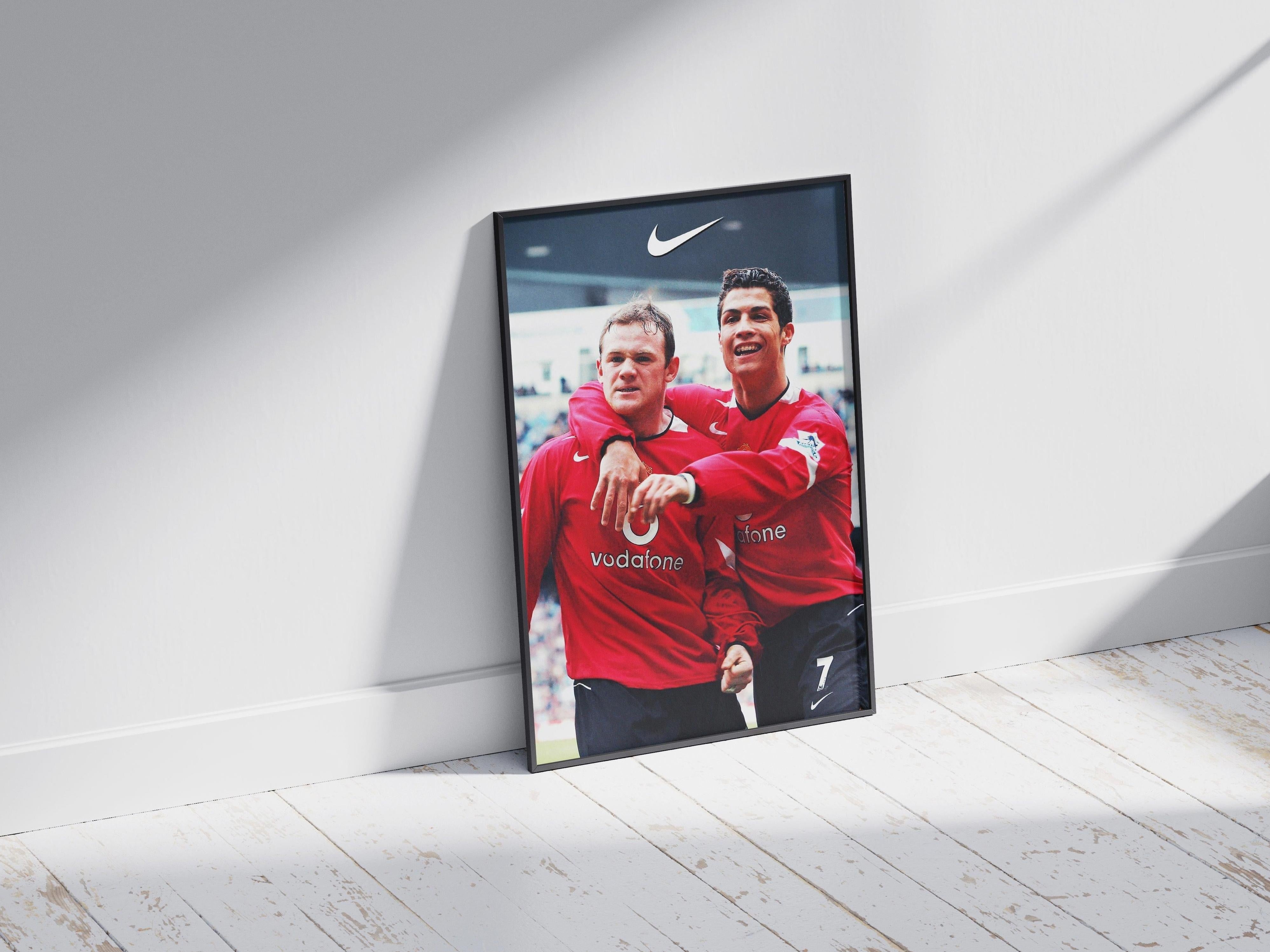 A framed Rooney x CR7 Poster by Retro1999 sits on a white wood floor, showing two soccer legends in red jerseys celebrating on the field beneath a Nike logo. Sunlight streams across the floor and wall.