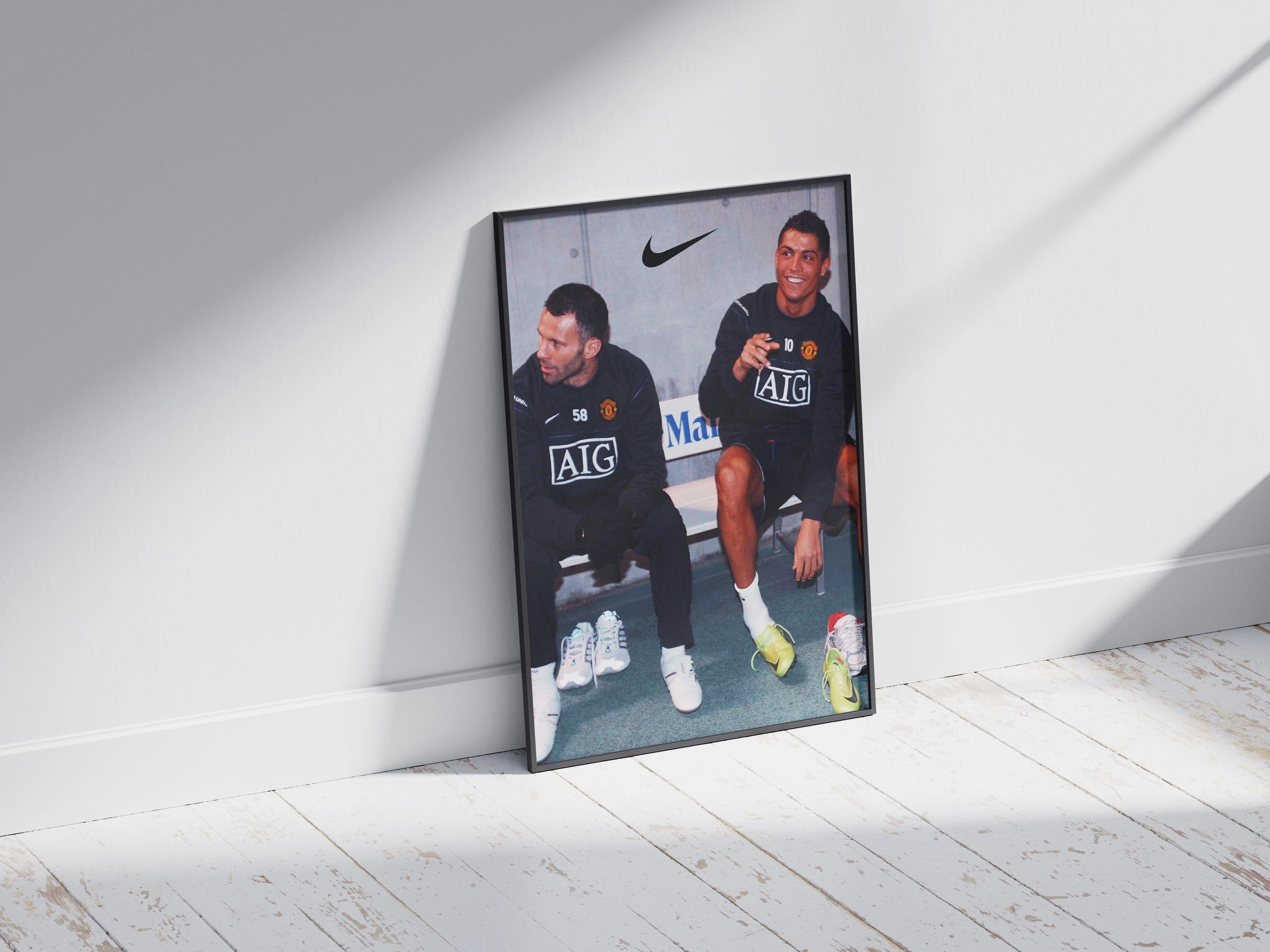 The Ryan Giggs x Cristiano Ronaldo Poster by Retro1999 shows both stars in black Manchester United training gear, sitting on a bench and smiling, with sunlight casting shadows across a white wooden floor.
