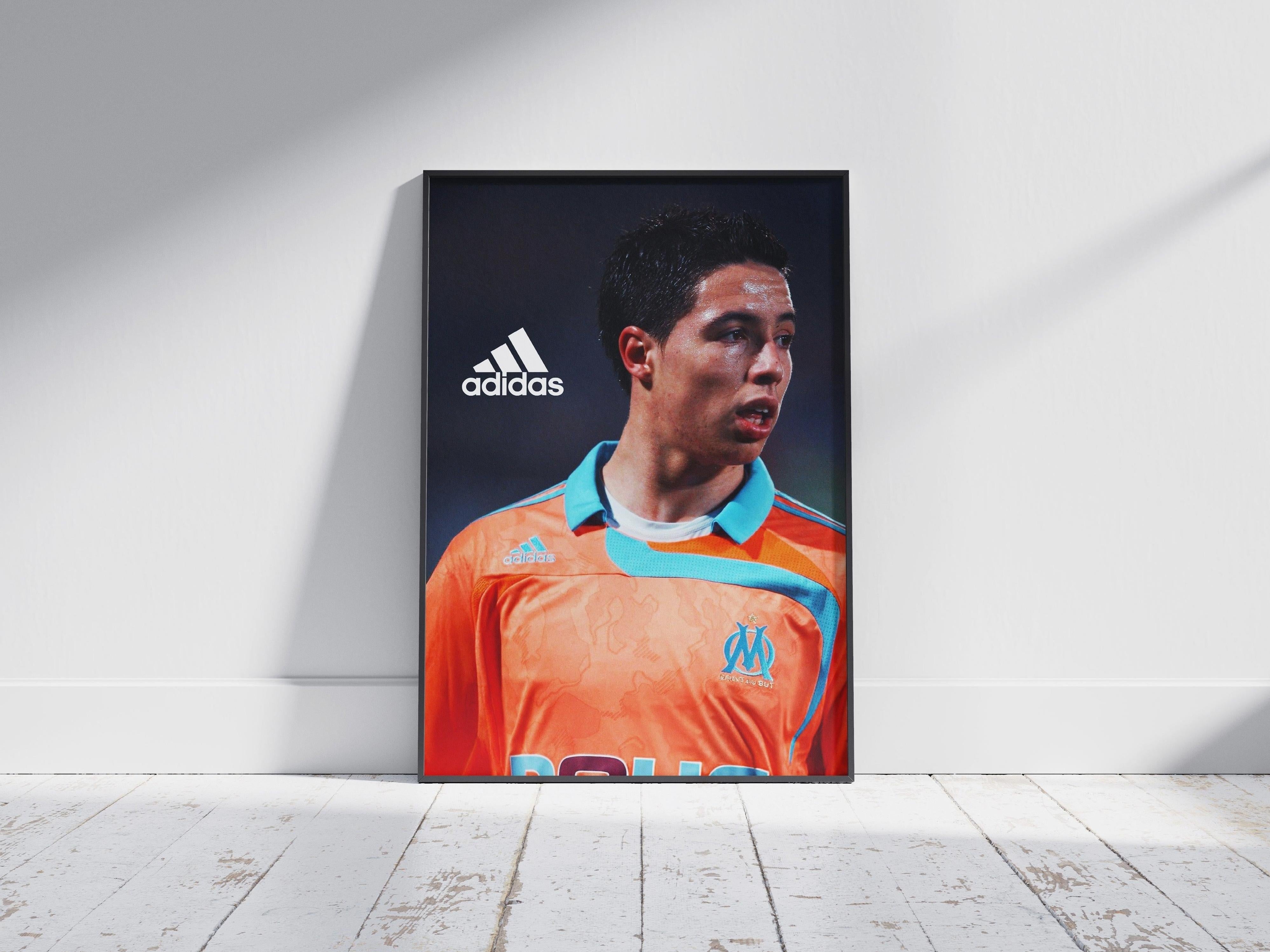 The Retro1999 Samir Nasri Marseille Poster features him in an orange and light blue jersey during a UEFA Cup match, displayed against a white wall on a wooden floor—a must-have for football memorabilia collectors.