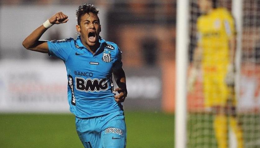 A soccer player in a blue Retro1999 Santos 2012-13 Third Shirt celebrates with a clenched fist and open mouth, running on the field, while a blurred yellow-clad goalkeeper stands by the goal in the background.