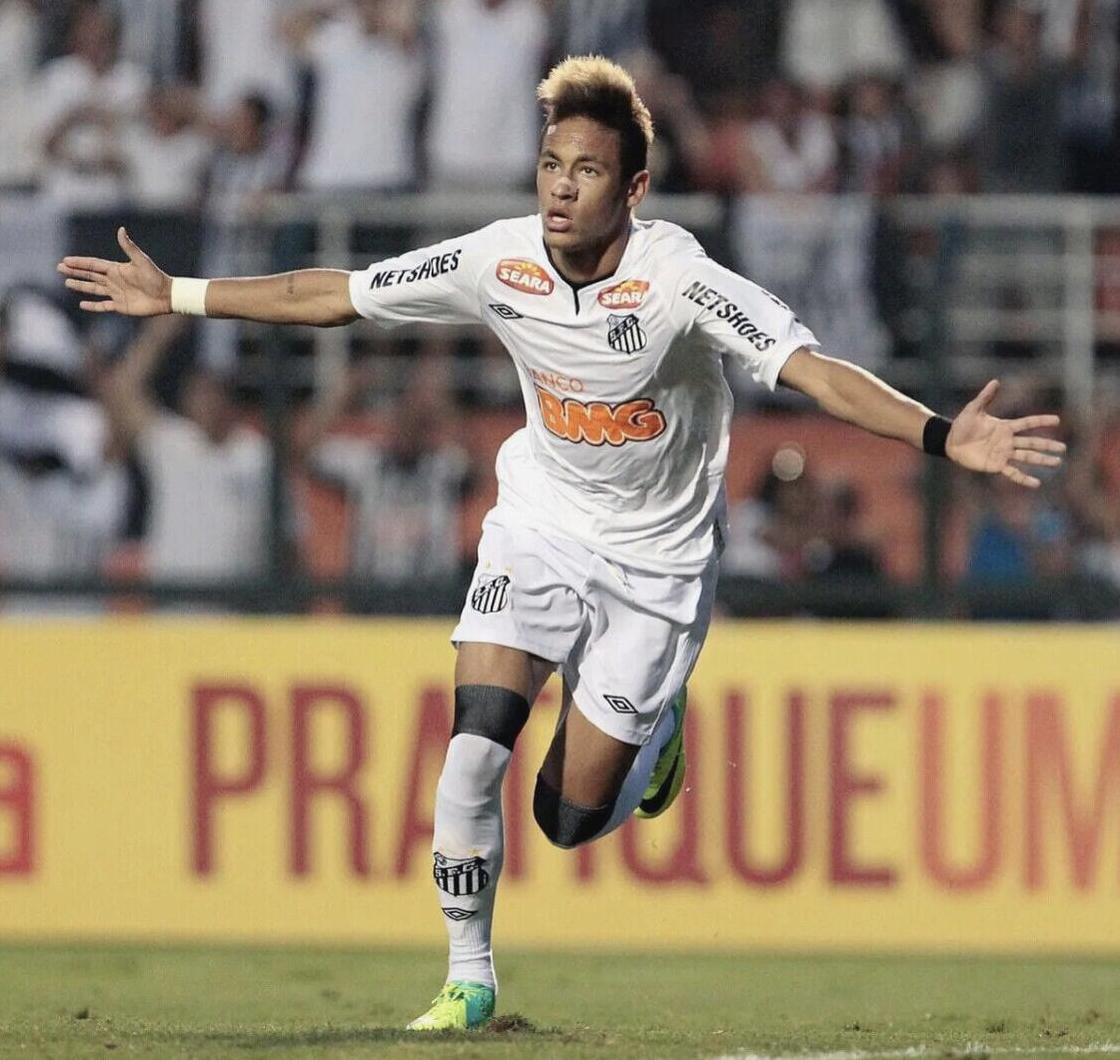 A soccer player in a Retro1999 Santos 2012-2013 Home Shirt celebrates on the field with arms outstretched, recalling Neymar’s iconic moments. Blurred spectators and a yellow ad board are visible in the background.