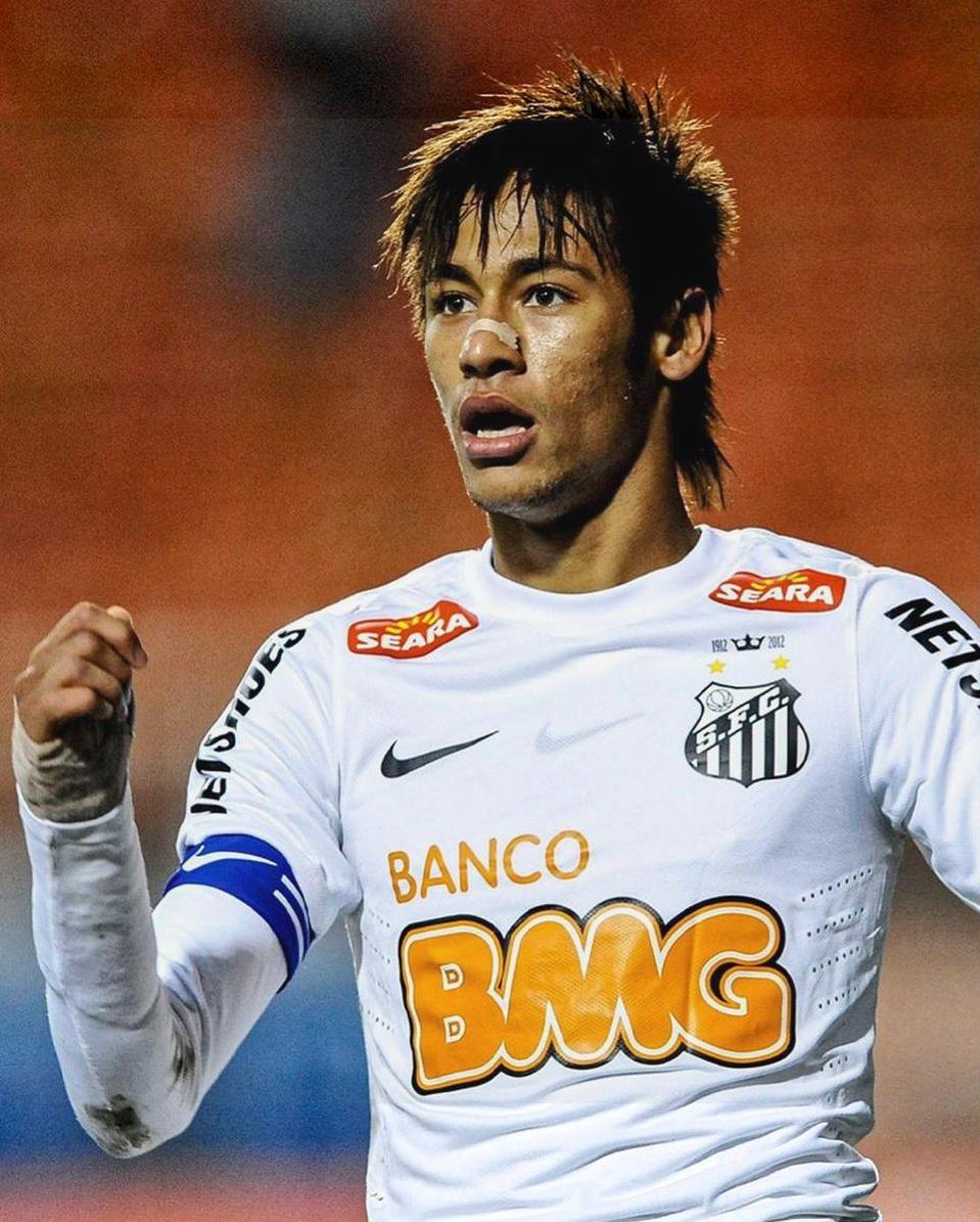 Neymar, with short brown hair and a blue captain’s armband, celebrates wearing the Retro1999 Santos 2012-2013 Home Shirt featuring the Banco BMG sponsor logo, with blurred orange stadium seats in the background.