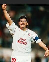 A soccer player celebrates in the Retro1999 Sevilla 1991-92 Home Shirt, featuring Super Nintendo on the front. Wearing a captain’s armband and number 10 shorts, he stands before a blurred La Liga stadium crowd.