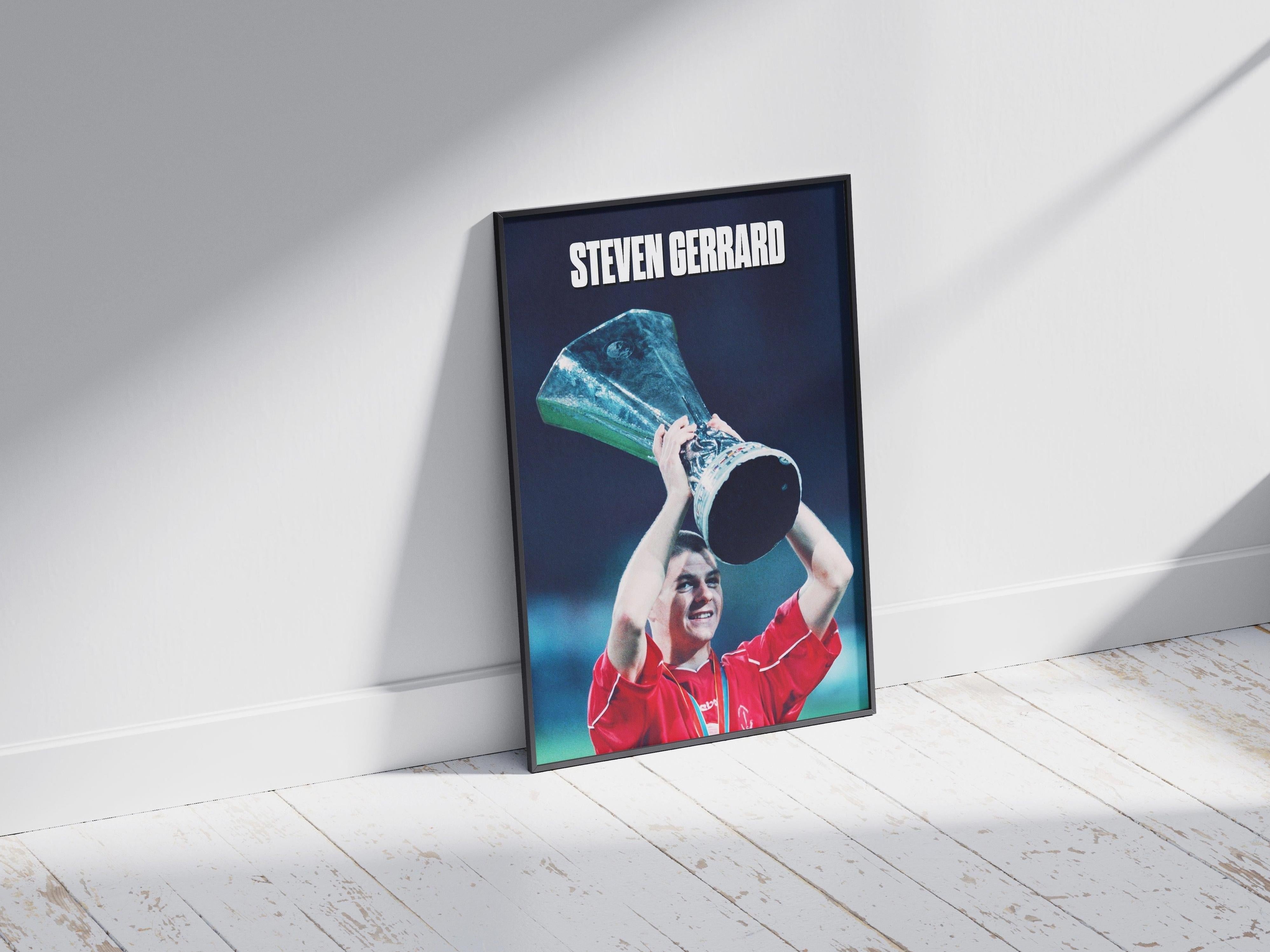 The Steven Gerrard x Champions League Poster by Retro1999 features a Liverpool FC player in a red jersey raising the Champions League trophy, with STEVEN GERRARD boldly displayed above, shown framed on a white wooden floor.