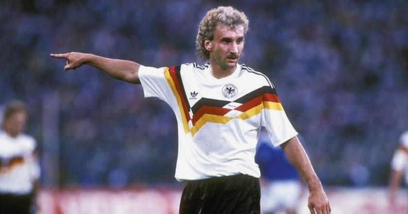 A soccer player in a Retro1999 West Germany 1990 Home Shirt with black, red, and yellow stripes points with his left arm during a match. He has curly blond hair and a mustache, with a blurred stadium and players in the background.