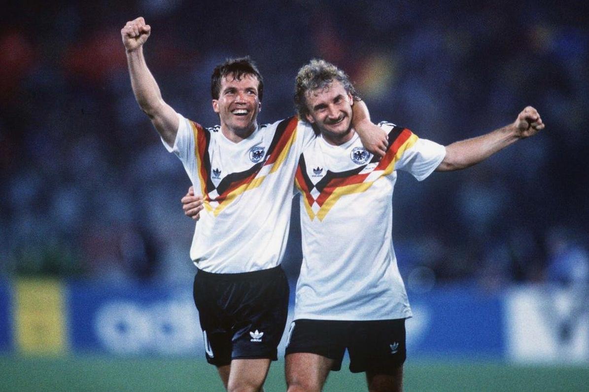 Two soccer players in Retro1999s West Germany 1990 Home Shirt celebrate a victory on the field, arms raised and smiling, under night stadium lights.